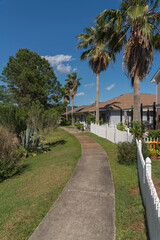 Fototapeta na wymiar Curved concrete walkway with bushes on the side outside the fenced residences in Navarre, Florida. Houses with palm trees inside their yard behind the picket fence near the curved path in the middle.