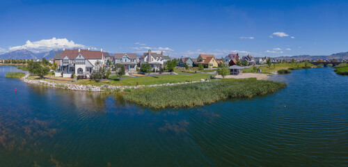 Fototapeta na wymiar Aerial view of lakefront single-family homes against the blue sky at Daybreak, Utah panorama. Lake with grasses near the shore and view of mountains at the background.