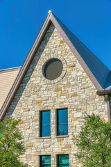 Vertical shot of a french country home with round attic window and reflective windows- Austin, Texas. There are trees outside near the reflective tall windows of the house with stone veneer sidings.