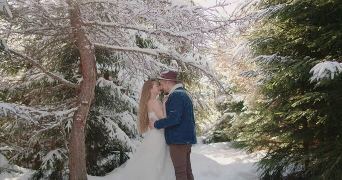 bride in knitted sweater wedding dress smiling laughing hugging kiss groom hat denim jacket under falling snow in forest Christmas tree. Slow motion 120 fps 4k. Snowing winter. blonde Caucasian woman