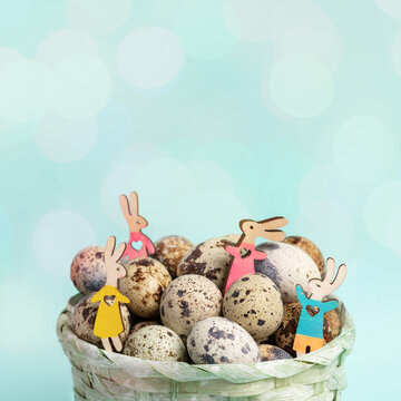 Four small wooden colored Easter bunnies among quail eggs in woven basket on pastel blue. Close up.