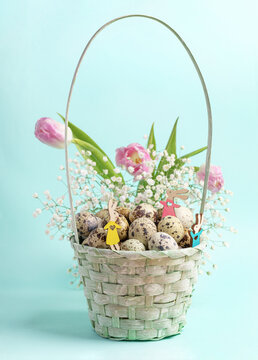 Small wooden Easter bunnies in blue woven basket with quail eggs, pink tulips, white gypsophila.
