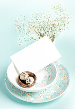 Easter card. Two quail eggs in nest, white empty note on plates with floral pattern on light blue.