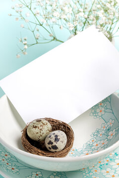 White blank note on plates with floral pattern, quail eggs in nest on light blue Copy space Close up