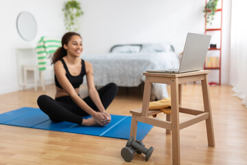 Smiling young sporty latin american woman sitting on bedroom floor doing online fitness exercise routine workout in her bedroom. Healthy lifestyle and sport concept. Focus on laptop