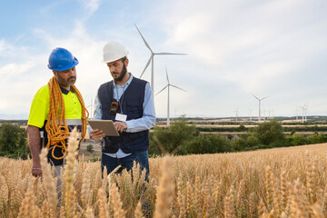 Electric engineer explaining maintenance worker plans for wind turbine farm using tablet. Copy space.