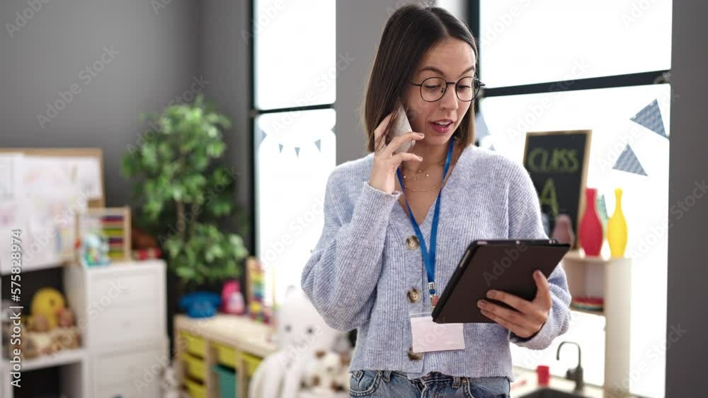 Wall mural Young beautiful hispanic woman working as a teacher talking on smartphone using touchpad at kindergarten