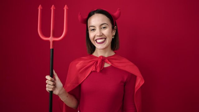 Young beautiful hispanic woman wearing devil costume holding trident over isolated red background