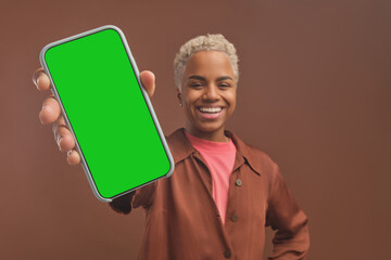 Young cheerful casual African American woman student holds out hand with phone to camera to show content in display with cool website or educational application stands in brown studio. Green screen