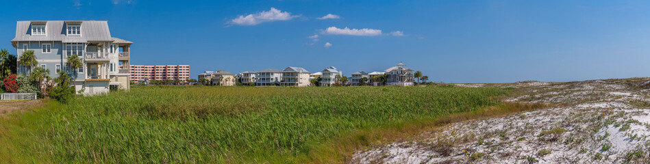 Fototapeta na wymiar Homes on the beach near the field of tall grasses and dunes at Destin Point in Destin, Florida. Panorama of a coastal residential area with three-storey homes and multi-storey apartment.