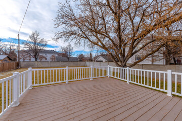Wooden deck with white railings in a backyard with grass, trees, and concrete wall fence. There is a leafless tree in the backyard and views of the neighborhood behind the wall under the cloudy sky.