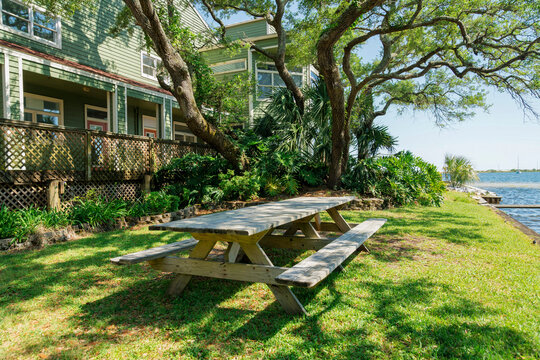 Navarre, Florida- Picnic area with wooden table and seats outside a fenced buildings near the ocean. Dining table under the tree with an ocean view on the right and buildings on the left.