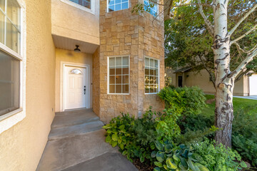 House entrance with plants and trees on the side heading to the white front door. There is a stucco wall on the left with window beside the entryway and bay windows with stone veneer on the right.
