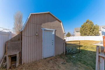 Shed with gambrel roof and white door at the backyard with white fence panel. There is a wood shed on the left near the fence and gate railings on the right access to the large yard with grass.