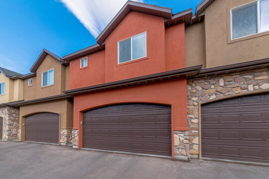 Townhouses with attached garages and painted orange and brown stucco and stone veneer walls. There are brown garage doors with arched entrances of two-storey townhomes under the clear sky.