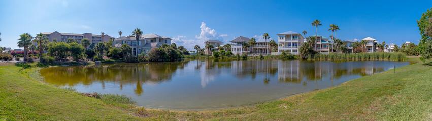 Fototapeta na wymiar Panorama of waterfront houses overlooking a freshwater lake in Destin Florida. Scenic residential landscape with homes, grassy field, water, and blue sky on a sunny day.