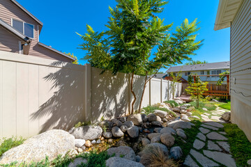 Side yard of a house with concrete pavers and small water stream in between rocks. There is a stream beside the beige fence on the left of the path beside the house with gray wood siding on the right.
