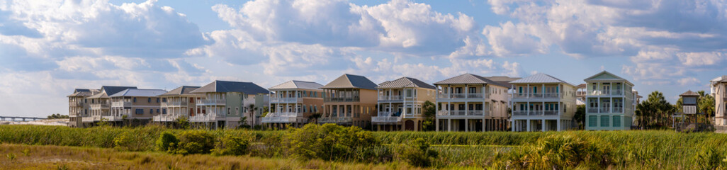 Fototapeta na wymiar Opulent multi storey houses with windows and terraces overlooking a lagoon. Panoramic landscape with lavish homes, lagoon, bright green foliage, and cloudy blue sky on a sunny day.