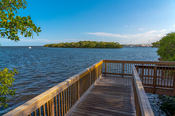 Boardwalk corner with railings and views of an island near the Dinner Key Marina at Miami, Florida. Corner of a wooden path near the trees with views of the water and island against the blue sky.