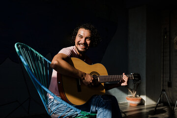 Young Hispanic man playing acoustic guitar sitting on floor in living room at Sunny Stylish Loft...