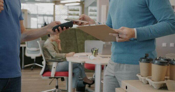 Close-up of businessman's hands paying for food delivery with smartphone indoors in office. Courier wearing uniform standing beside.