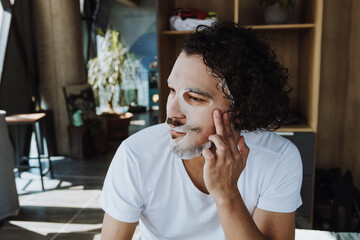 hispanic Man applying exfoliating facial mask treatment taking care of skin, morning face wash routine for cleaning in loft apartment in Mexico Latin America