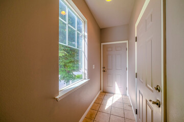 Interior of a house with narrow hallway leading to rooms with closed white doors. Lush green trees can be seen from the window on this sunny day.