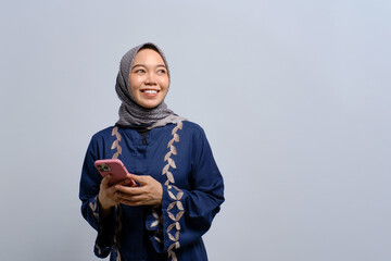Smiling young Asian Muslim woman using mobile phone and looking away at copy space isolated over white background