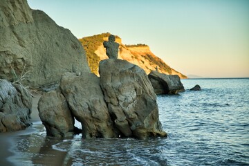 View of a beach of Corfu in Greece along cliffs in the foreground rocks with a stone figure in the water. - obrazy, fototapety, plakaty