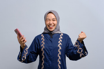 Excited young Asian Muslim woman using mobile phone and celebrating success, getting good news isolated over white background