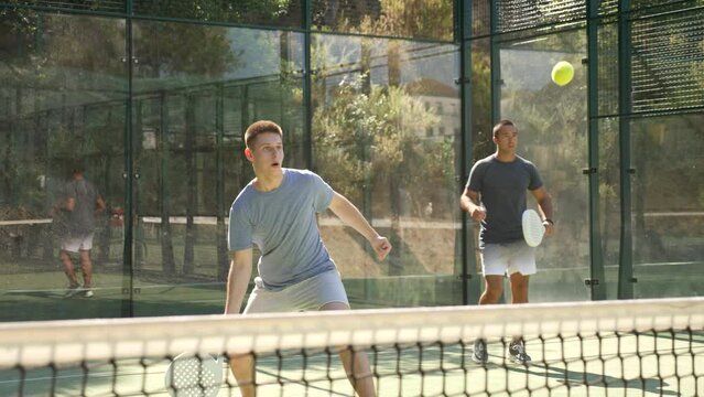 Portrait of emotional determined young guy playing padel tennis on open court in summer, swinging racket to return ball over net. Sportsman ready to hit volley