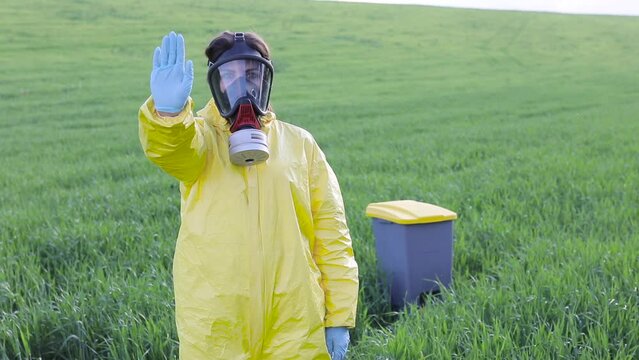 A woman in a yellow protective suit stands in the middle of a green field in a protective gas mask and shows a gesture to call to stop and save the planet.