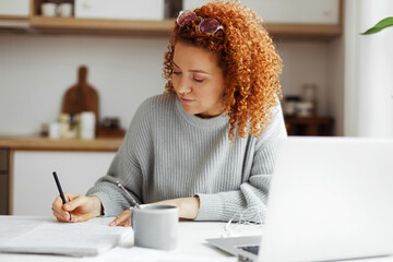 Redhead female interpreter freelancer with curly hair working from home, writing translation in her copybook sitting at kitchen table in front of laptop, translating article for web site - obrazy, fototapety, plakaty
