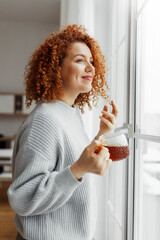 Vertical portrait of gorgeous redhead curly female in sweater drinking hot tea to warm up admiring beauty of snowy winter standing next to big window and looking through touching her hair - 575851179