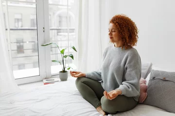 Abwaschbare Fototapete Young plus-size redhead woman with fluffy curly hair sitting in lotus posture on bed near big window keeping hands on knees, meditating, trying to relax her mind and relieve stress © shurkin_son