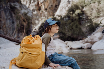 School boy with yellow backpack sits on a riverside rock in the canyon with mountain cliffs in the...