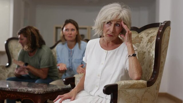 Dissatisfied mother-in-law siting on a chair against of a confused husband and wife on the sofa in the living room at home
