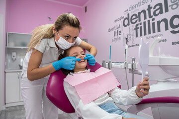 female dentist shows the treated teeth to the little girl patient in front of the mirror.