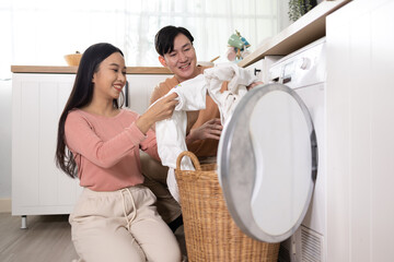 Young Asian married happy couple smiling and doing laundry at home. Boyfriend and girlfriend putting clothes in front loading washing machine together