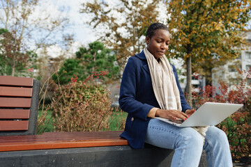 Fototapeta premium Young african businesswoman using laptop while sitting on the bench in the city