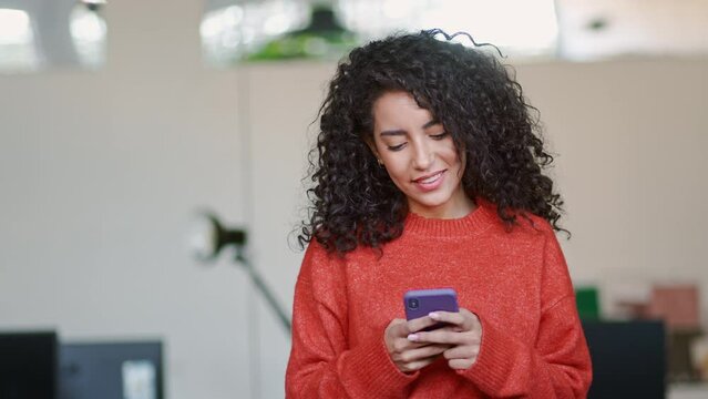 Smiling young professional latin business woman, happy lady corporate leader holding cellular phone working standing in modern office using mobile apps cellphone technology device looking at cell.
