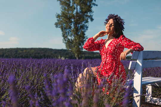 A Young Woman In A Wreath On Her Head Sits On A White Bench In A Lavender Field