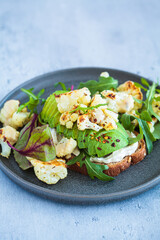 Vegan toast with avocado, baked cauliflower, lettuce and tahini on dark plate. Healthy plant based organic diet.
