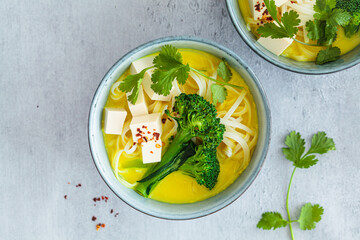 Vegan laksa with rice noodles, broccoli and tofu in blue bowls, gray background.