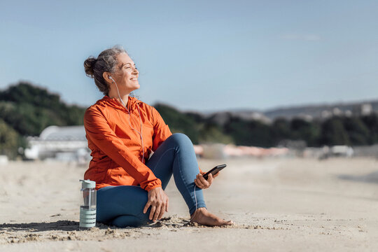 Smiling woman listening to music through in-ear headphones at beach - Powered by Adobe