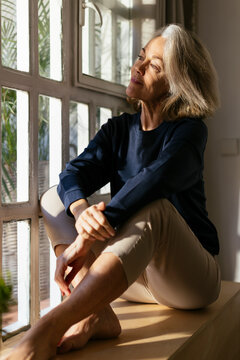 Thoughtful mature woman sitting on window sill at home