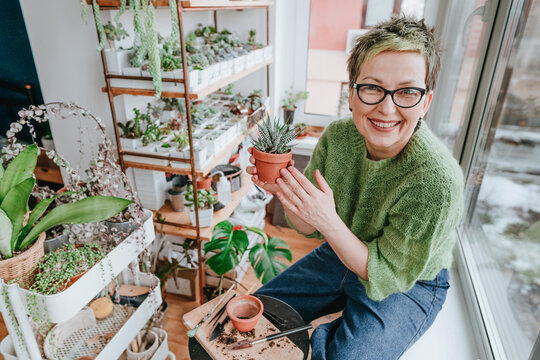 Happy woman holding potted plant sitting on window sill at home - Powered by Adobe