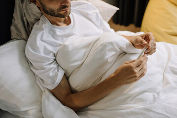 a man lies covered in bed, holding a thermometer in his hands. conceptual photo of a disease or disease
