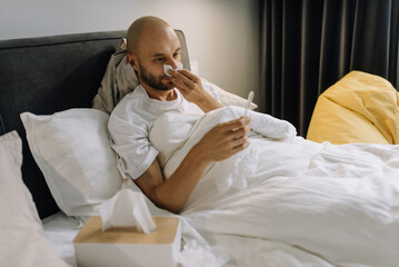 A sick man lies in bed and blows his nose with a paper napkin while holding a thermometer