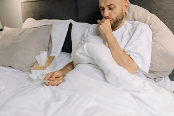 A man covers his mouth with his hand while coughing and holds a thermometer in his other hand while lying on the bed in the bedroom. His facial expression expresses concern and discomfort.
